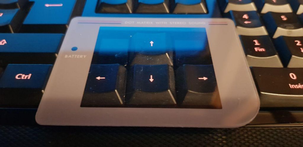 A Game Boy screen cover laying on top of the arrow keys on a red-lit computer keyboard.